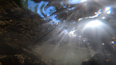 Flowing water, skylight of sun through water, stones lying at bottom, underside of surface of water stream. Sun glare moves over surface of stones. Nature backdrop, natural background. Environment