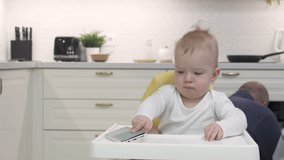 Father handing mobile device to child to entertain him, baby boy holding mobile phone sitting on high chair in the kitchen. High quality 4k footage