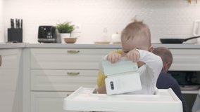 Father handing mobile device to child to entertain him, baby boy holding mobile phone sitting on high chair in the kitchen. High quality 4k footage
