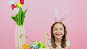 Close up video of an adorable girl wearing bunny ears on head sitting behind the chair with flowers in vase and basket waving with a hand, Easter holiday concept.