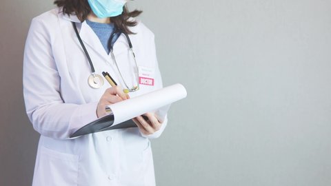 female doctor in white uniform, stethoscope around neck, protective facial mask, filling papers on black clipboard. unrecognizable caucasian woman physician pen in hand, writing prescriptions, medical