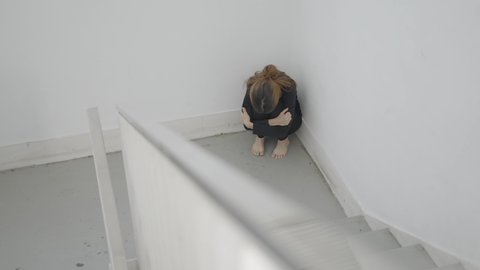 Young scared woman sits curled up in corner of stairwell, from above