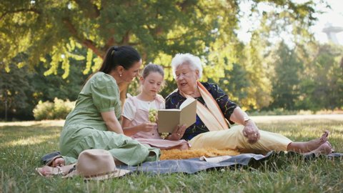 Small girl with mother and grandmother resting in a park, reading a book.