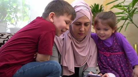 Happy Eid Celebrations Online. Traditional Muslim family, mother in hijab and children together at home using smartphone to call friends