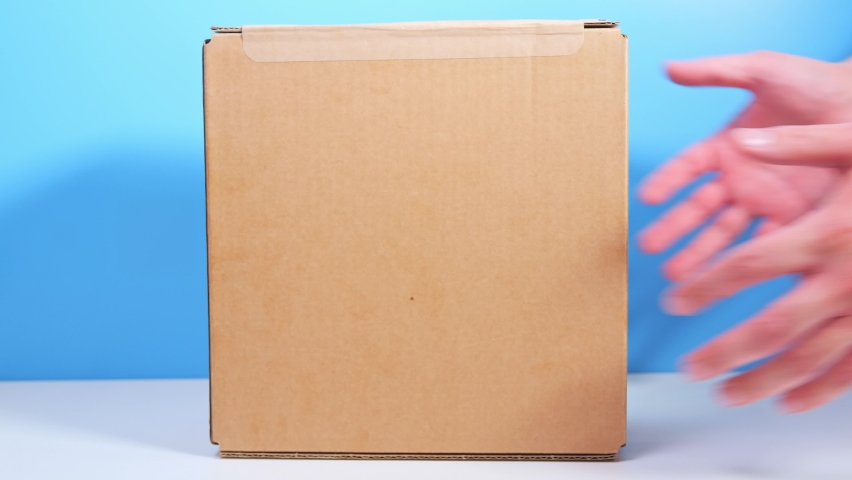 Courier girl passes cardboard box to buyer. Express delivery of high-tech equipment. Postal or courier service. Cardboard box on a white table and a blue wall background. | Shutterstock HD Video #1088273451