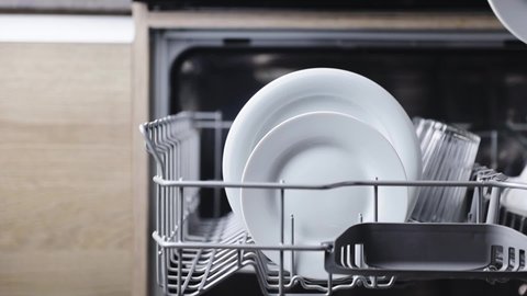 Close-up of woman hand takes a washed plate from the basket of an open automatic stainless built-in dishwasher machine with clean utensils inside in modern home kitchen. Household domestic life