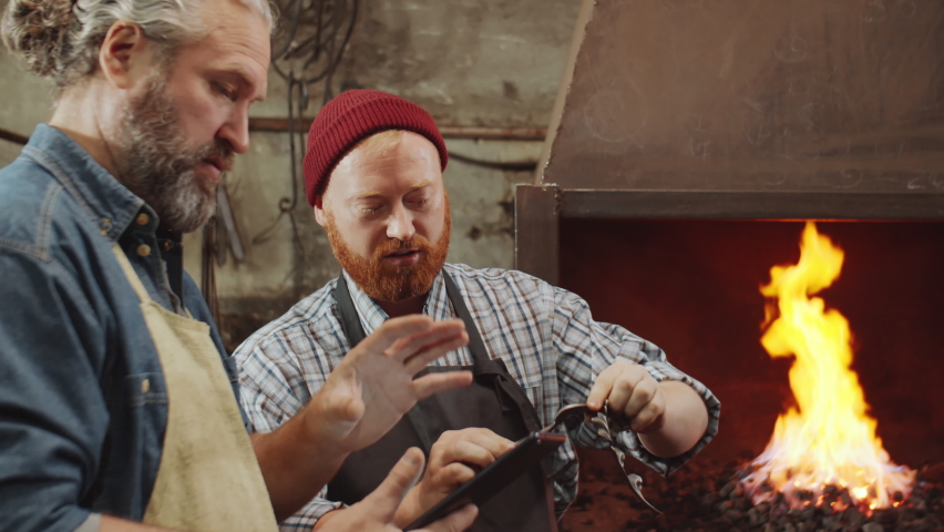 Two professional blacksmiths discussing metal details and working on digital tablet in smithy