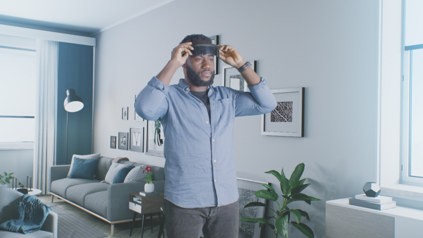 African American man putting on VR headset and looking around with smile while exploring futuristic cyberspace of metaverse in living room at home Royalty-Free Stock Footage #1088275535