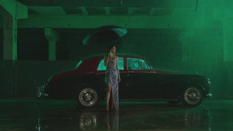 Beautiful Elegant Young Woman Standing and holding umbrella near old retro classic car. Girl with umbrella stands in street under the rain . Old fashioned luxury style concept . Raining outside 
