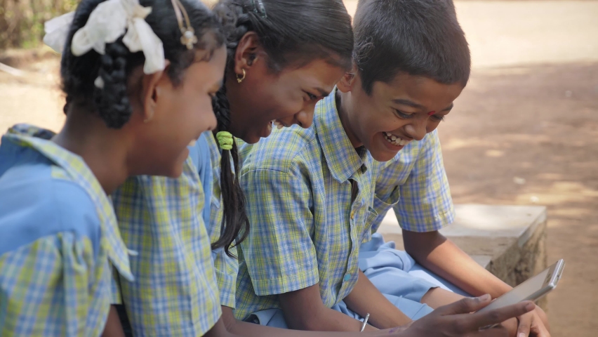 Cheerful smiling Indian group of young Rural school children laughing in uniform sitting together and watching online educational tutorials or videos on a mobile phone or digital Tablet in daylight. | Shutterstock HD Video #1088278305