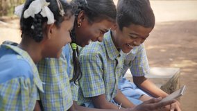Cheerful smiling Indian group of young Rural school children laughing in uniform sitting together and watching online educational tutorials or videos on a mobile phone or digital Tablet in daylight.