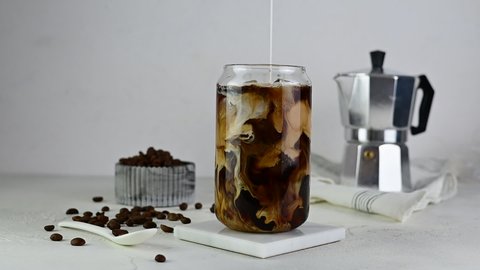 A glass glass with ice and black coffee on a marble stand. Milk pours into a glass. A coffee pot and coffee beans in a vase in the background. Light table
