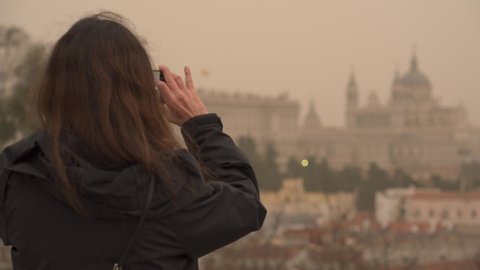 MADRID, SPAIN - March 15 2022. A woman at a viewpoint takes a picture with her smartphone of Madrid’s Royal Palace and Almudena Cathedral amid an orange sky due to a dust storm from the Sahara desert 