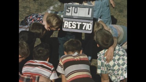 1940s: Field, boys huddle in circle. Boy sits on folding chair, holds arm up, pinches muscles, turns, looks over shoulder, grabs rolling tray table.