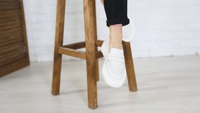 Presentation of white shoes with plain massive soles. Lady in modern footwear sits on the tabouret waving her feet. Close up.