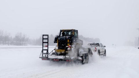 BAXTER, MN - 22 FEB 2022: White pickup towing a trailer with a Deere Track Loader with dozer blade during a winter storm in Minnesota, while snow falls and wind blows.