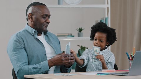 African american family sitting at desk adult father home teacher teaches financial literacy arithmetic little daughter girl learns count with help paper money dollar bills joyful child plays with dad
