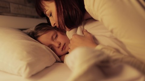 Sweet dreams. Cute little child girl sleeps in the bed, while her mommy adjusting the blanket and kissing her chick. Sleep well concept. Mom and daughter going to sleep
