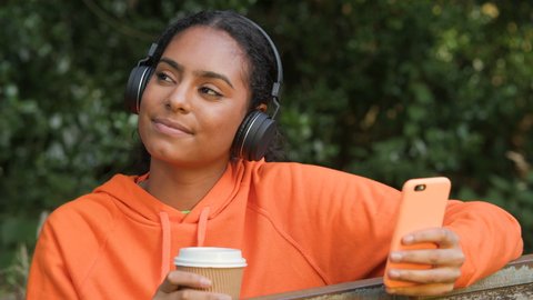 Beautiful African American girl teenager young woman listening to music on her smartphone or cell phone wearing wireless headphones and drinking take out coffee