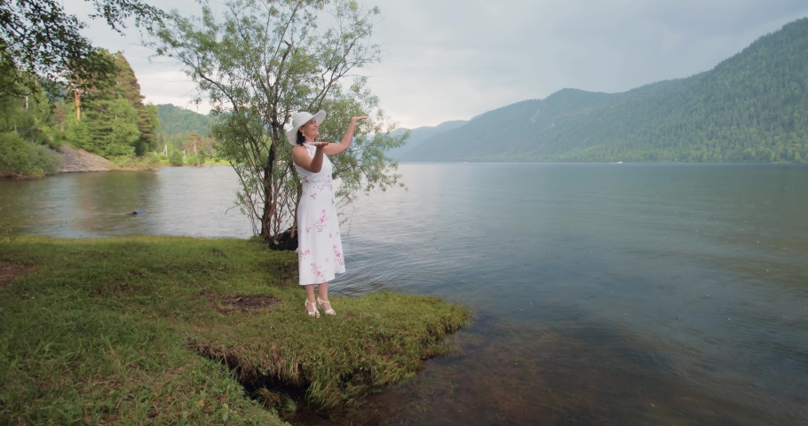A beautiful and attractive lady in a white dress and hat stands on the shore of a mountain lake and rejoices at the rain that has begun. She raised her hands up and catches raindrops with her palms.