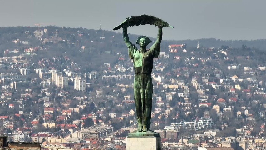 Beautiful green Freedom and liberty statue in the center of Budapest with a densely built neighborhood on top of the green hills in the background. Drone close up lifting tilt down shot