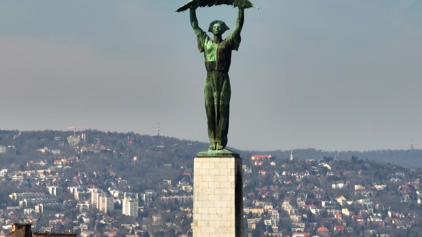 Green statue for freedom in the center of Budapest in Hungary with densely populated residential areas on top of a hill in the background. Close up lifting drone shot