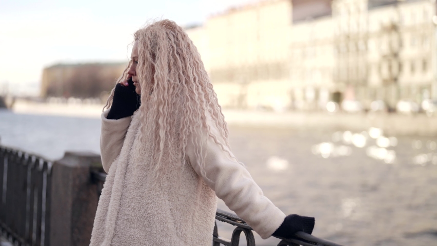 An adult woman is having an unpleasant conversation on a mobile phone on the street near the river. Close-up of an excited girl with long hair is surprised during a phone call.