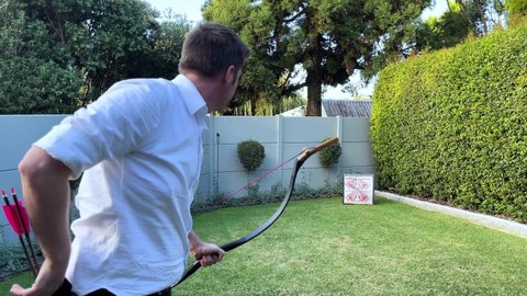 Caucasian man in 30's shooting arrows a Tibetan horse bow into a square target in a suburban backyard in the late afternoon