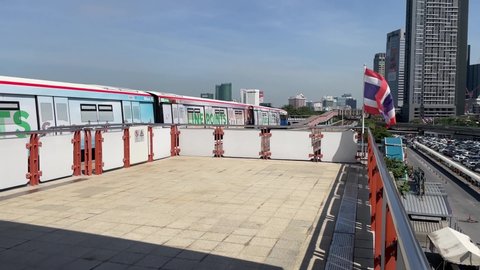 BANGKOK, THAILAND - Circa November, 2021: BTS Skytrain Public Transport departing Mo Chit Station, interchange station to Chatuchak MRT subway. Beautiful skyline and Thai flag waving - POV shot 60fps