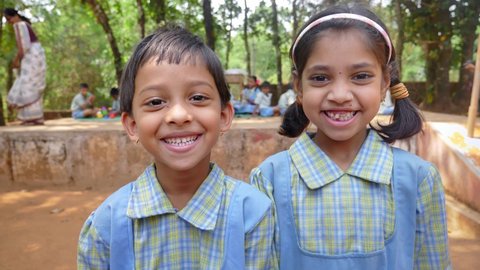Two cute little smiling Indian Rural schoolgirls in uniform looking at the camera with a group of students studying in the background in a village area. concept of female education and literacy