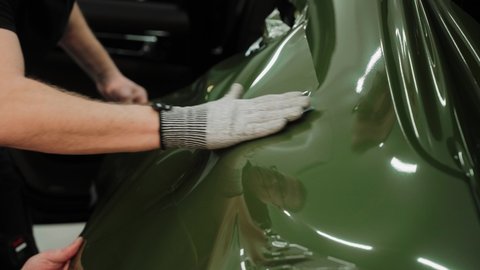 Slow motion shot of man vinyl wrapping the car, smoothing the wrap with his hands. Process of wrapping a car in a garage. Dark green vinyl car wrap. High quality 4k footage