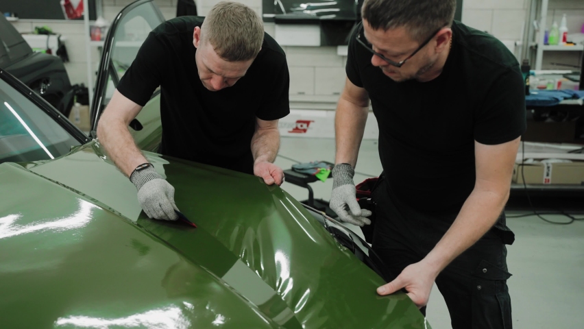 Two men are vinyl wrapping a car in dark green color using plastic cards. Process of vinyl wrapping a car hood in a car studio. Dark green car vinyl wrap. High quality 4k footage Royalty-Free Stock Footage #1088310813