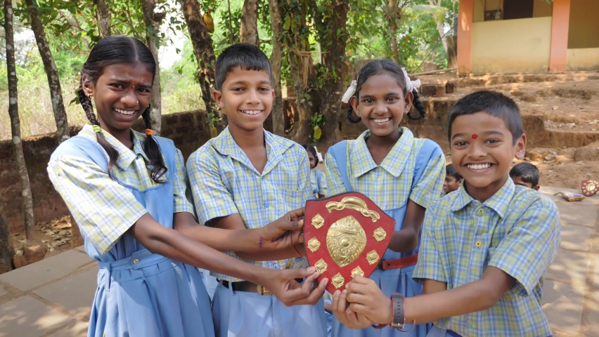Group of cheerful smiling young Indian rural primary school male and female children in uniform together holding a prize shield or award after winning a competition standing in the village premises.  Royalty-Free Stock Footage #1088311067