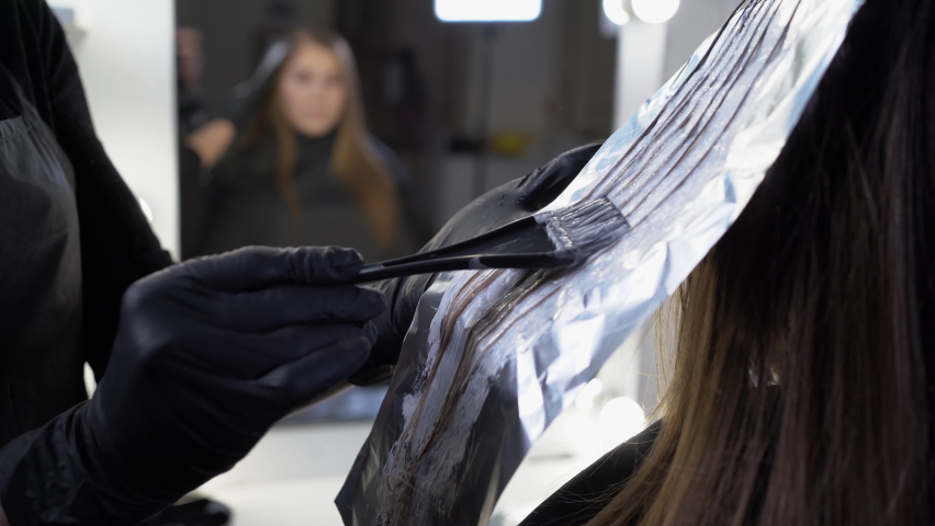Close-up side view of a strand of female hair on a special sheet of foil, the master applies hair dye with a brush. The process of highlighting by a professional master in a beauty salon. Life style | Shutterstock HD Video #1088311193