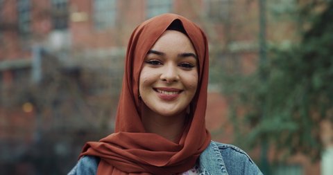 Cinematic shot of young happy Arabian muslim woman is adjusting her hijab and smiling  in camera outdoors.