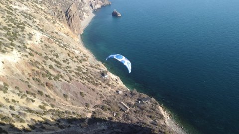 Aerial drone view of a man flying a white and blue paraglider over a hill and trees to the sea waves near the rocks. Active paraglider flight over the seascape with clear skies at suset. Extreme sport