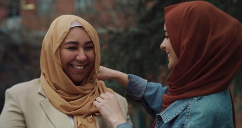 Cinematic shot of young happy Arabian muslim woman is adjusting hijab to her friend while having fun to pass time together outdoors.