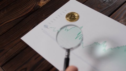 A graph of the growth of cryptocurrency in a magnifying glass, in the background lies a gold coin Bitcoin. Digital money, banking, investment, finance and business concept.