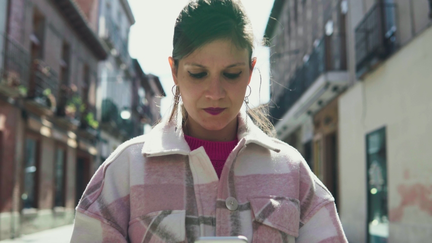 Brunette woman dressing youthfully talking to her phone while waiting in a central alley of the city and looking at the camera.Front image of a young girl wearing a shirt and holding a white cellular.
