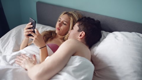 Nice girl staring mobile phone in bed. Sad woman and man, husband in bed, lack of sexual activity in family. Young unhappy couple problem in bedroom, quarrel between lovers. Divorce, marriage