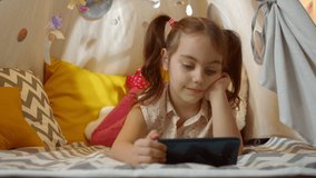 Preschool girl is watching a cartoon on her smartphone while lying in a tent at home. Preschooler is playing on a mobile phone. Child uses the phone for games. Child plays a video game at home