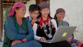 A cheerful Asian Indian group of rural family members including mixed aged women wearing traditional costumes and a kid smiling and watching funny online movie or video on a laptop in a remote village