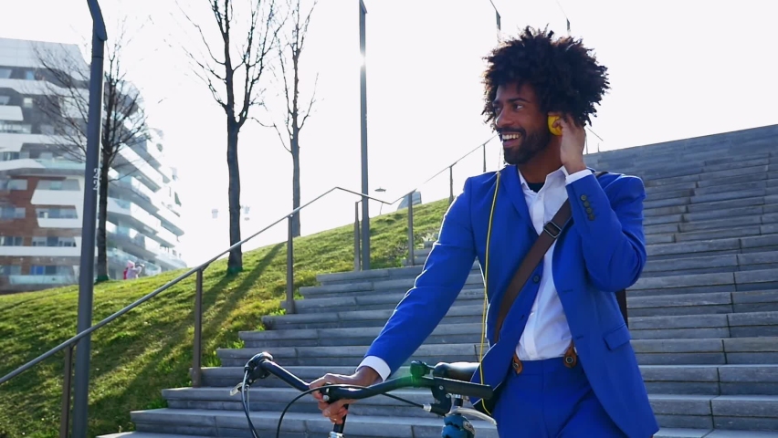 Handsome latin adult man wearing electric blue smart casual suit walking with bicycle and listening music with headphones - Hispanic cool business man with curly hairstyle strolling in the city