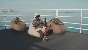 Wide shot of young Black man sitting on bean chair on pier on lake in summer, smiling and waving hand on smartphone camera, video calling