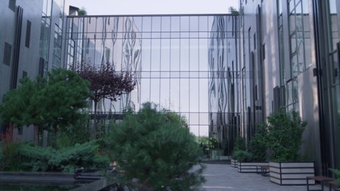 Neat garden at glass building in modern downtown district. Empty peaceful recreation area lush green trees at office construction. Large panorama windows reflecting clear sky. Architecture concept.