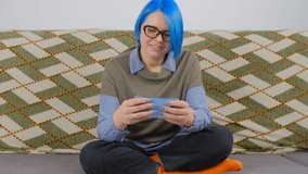 Mobile gamer woman playing game app on smartphone. Cute white female with dyed blue hair wearing nerdy glasses plays modern gamers on modern mobile phone with happy smile while sitting on couch