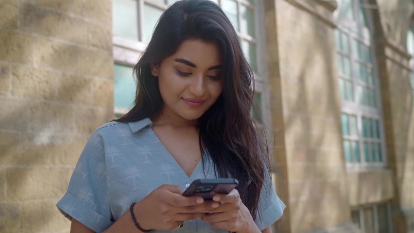 A young Modern happy Indian Asian hipster modern stylish female or woman is standing outdoors against an old vintage downtown building and chatting or typing a text message on a Mobile phone.  Royalty-Free Stock Footage #1088330561