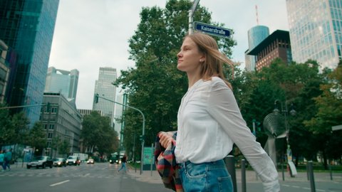 Millennial European Woman in White Shirt Crosses the Road Walking in City Downtown Streets in Frankfurt am Main, Germany at Daytime. Urban Lifestyle and Business concept. 4K low angle tracking shot