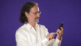 Portrait of happy mature businessman having video chat using mobile cell phone isolated on purple background in studio with copy space.