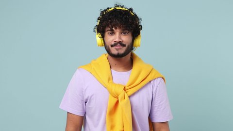 Vivid young bearded Indian man 20s wears violet t-shirt listening music in headphones dance sing song have fun gesticulating hands enjoy isolated on plain pastel light blue background studio portrait